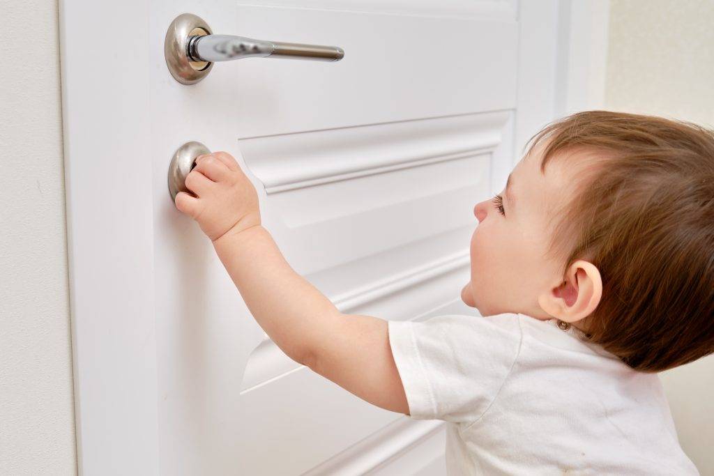 Toddler baby opens the lock, child hand close-up. White wooden door, metal door handle and baby hand